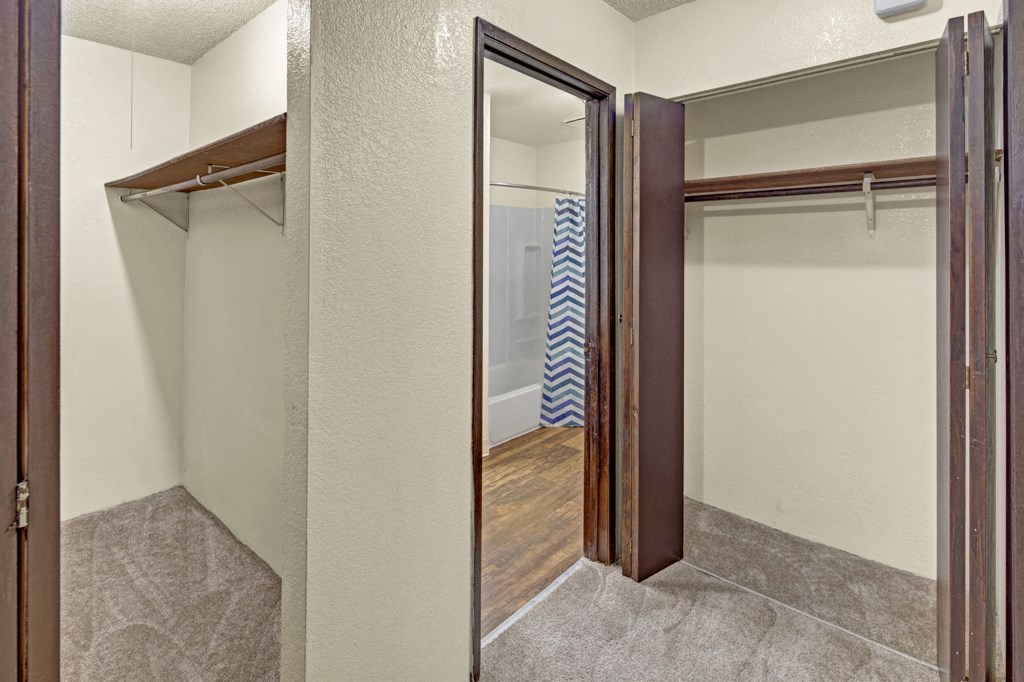 A small, empty walk-in closet here at Hillside Chalet Apartments with beige walls, open wooden shelves, and brown carpet. Leads to a bathroom with a chevron-patterned shower curtain.