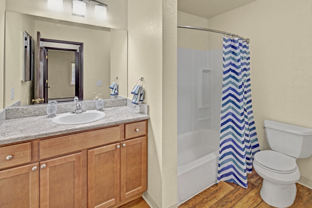 Bathroom here at Hillside Chalet Apartments with wooden cabinets, granite countertop, and mirror above a sink. A shower with a blue chevron curtain and a toilet beside it. Cozy ambiance.