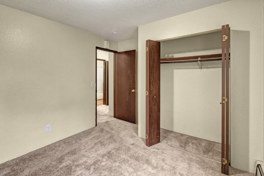 Empty bedroom here at Hillside Chalet Apartments with textured beige walls and carpeted floor. An open closet with wooden doors is on the right, and a doorway leads to a bathroom.