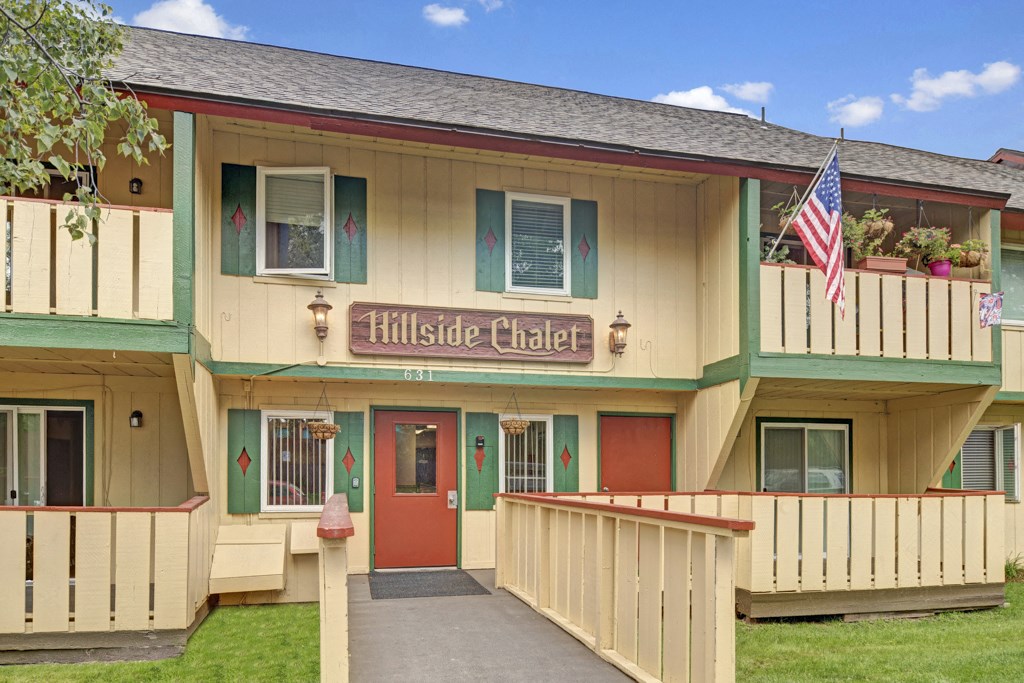 Two-story Hillside Chalet Apartment building with a rustic design, labeled "Hillside Chalet." Features balconies with potted plants, a wooden fence, and an American flag.