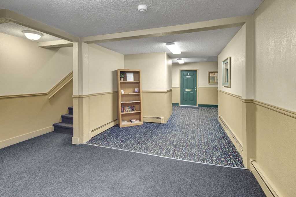 A quiet, well-lit hallway here at Hillside Chalet Apartments with cream-colored walls and patterned carpet. A green door is at the end, with stairs and a bookshelf on the left.