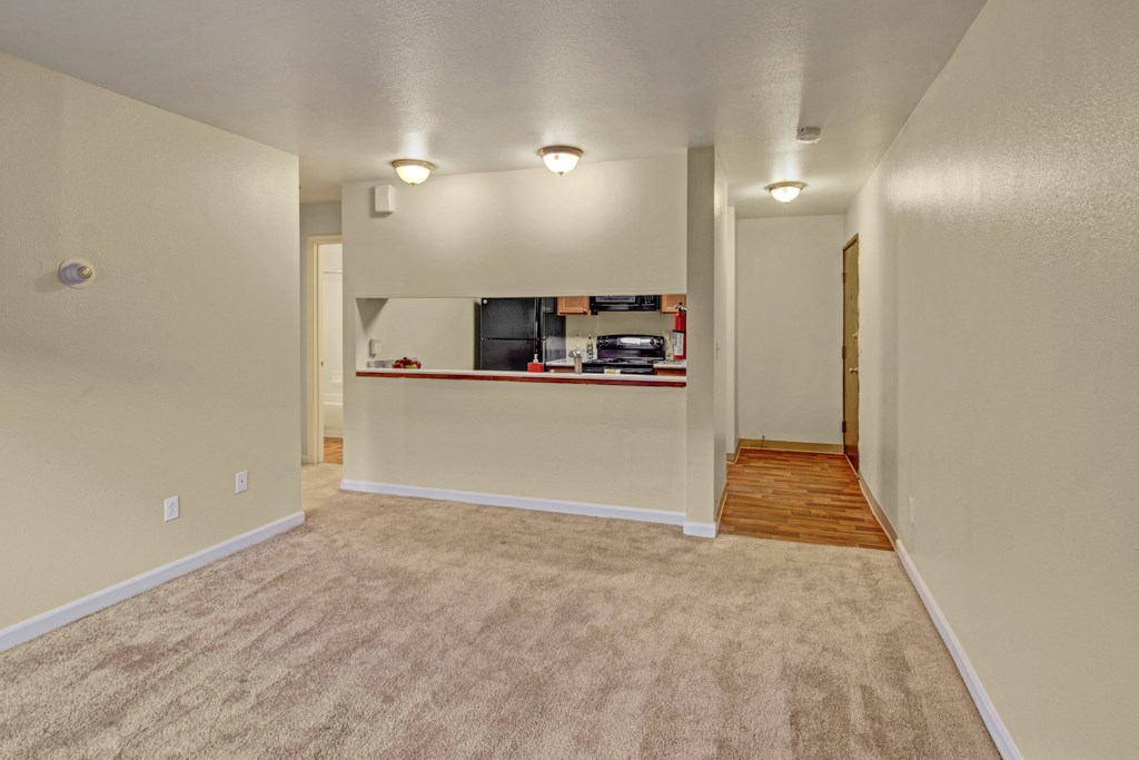 Quiet apartment interior here at Hillside Chalet Apartments with beige carpeted living area leading to a small kitchen. Warm lighting creates a cozy and inviting atmosphere.