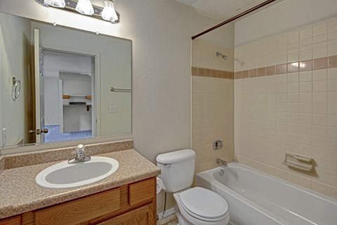 A bathroom here at La Maisonnette Apartments with beige tiled walls features a bathtub, a white toilet, and a brown vanity with a sink. A mirror reflects a doorway to another room.