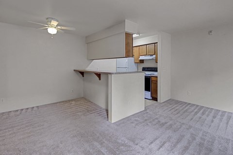 Simple apartment living area here at La Maisonnette Apartments with beige carpet, small kitchen with white appliances, wooden cabinets, and a ceiling fan. Bright and minimalistic.