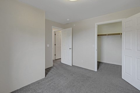 Empty bedroom here at La Maisonnette Apartments with gray carpet and light beige walls. Two open doors reveal a closet and a hallway. The space feels clean and minimalistic.