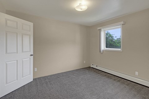 Bedroom here at La Maisonnette Apartments with beige walls, a gray carpet, and a white door ajar. A window with blinds lets natural light in, creating a calm, neutral ambiance.