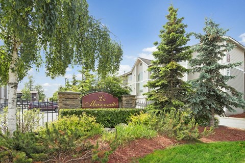 Lush garden with trees surrounds a sign labeled "La Maisonnette." A light-colored building is in the background under a clear blue sky.