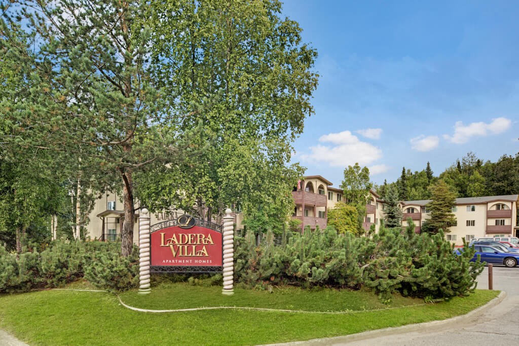 A serene apartment complex with a sign reading "Ladera Villa" amidst lush greenery and tall trees. The buildings are cream with arched windows. The sky is clear.