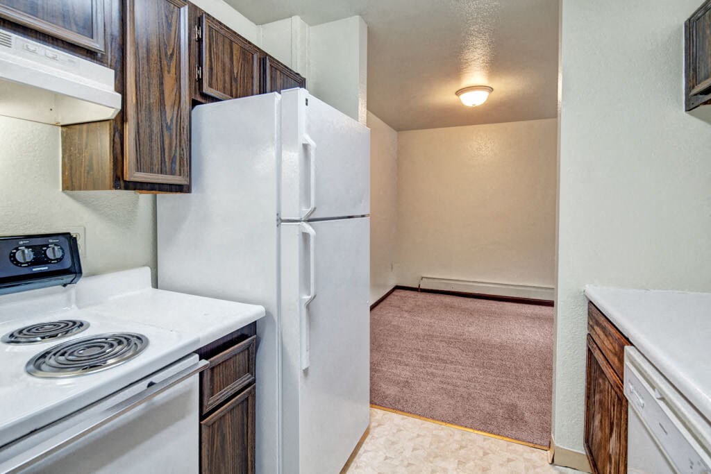 Compact kitchen here at Ladera Villas Apartments with wooden cabinets, white stove and fridge. Leads to a carpeted dining area, softly lit by a round ceiling light. Cozy and simple.