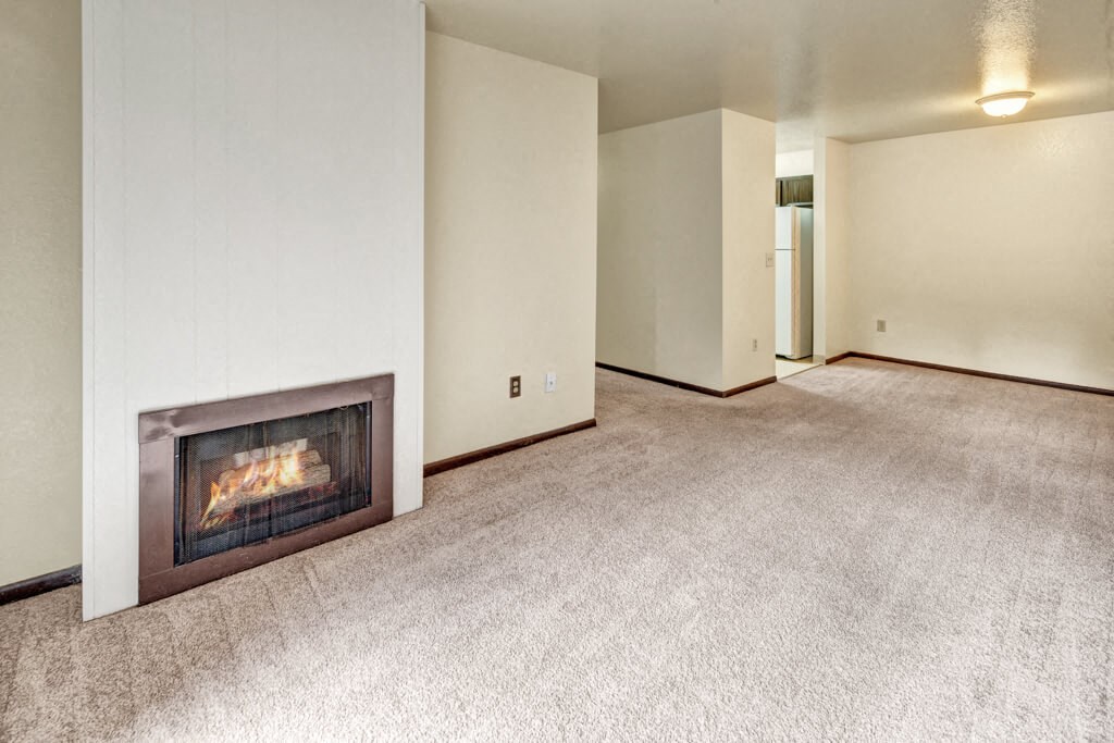 Cozy living room here at Ladera Villas Apartments with beige carpet and soft lighting. A corner fireplace adds warmth. A glimpse of the kitchen area is visible through an open doorway.