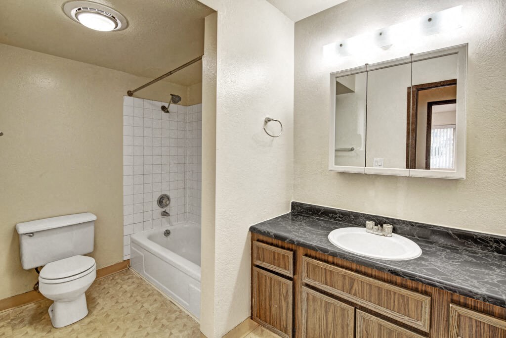 A tidy bathroom here at Ladera Villas Apartments with a bathtub-shower combo, white tiles, and a white toilet. A wooden vanity with a marble countertop sits beneath a lit mirror.