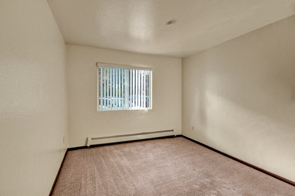 Bedroom here at Ladera Villas Apartments with beige carpet and cream walls, lit by sunlight through vertical blinds on a window. The tone is warm and inviting.