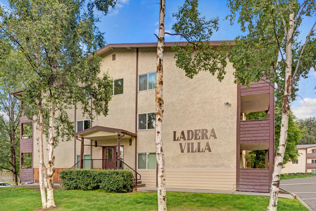Three-story beige Ladera Villa apartment building labeled "Ladera Villa" with brown accents. Surrounded by lush green trees and grass under a clear blue sky.