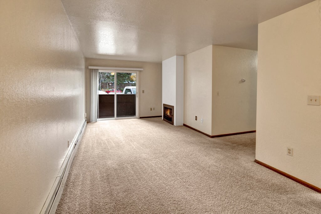 A spacious, empty living room here at Ladera Villas Apartments with beige carpet and white walls. A lit fireplace adds warmth. A sliding glass door opens to a patio, revealing parked cars outside.