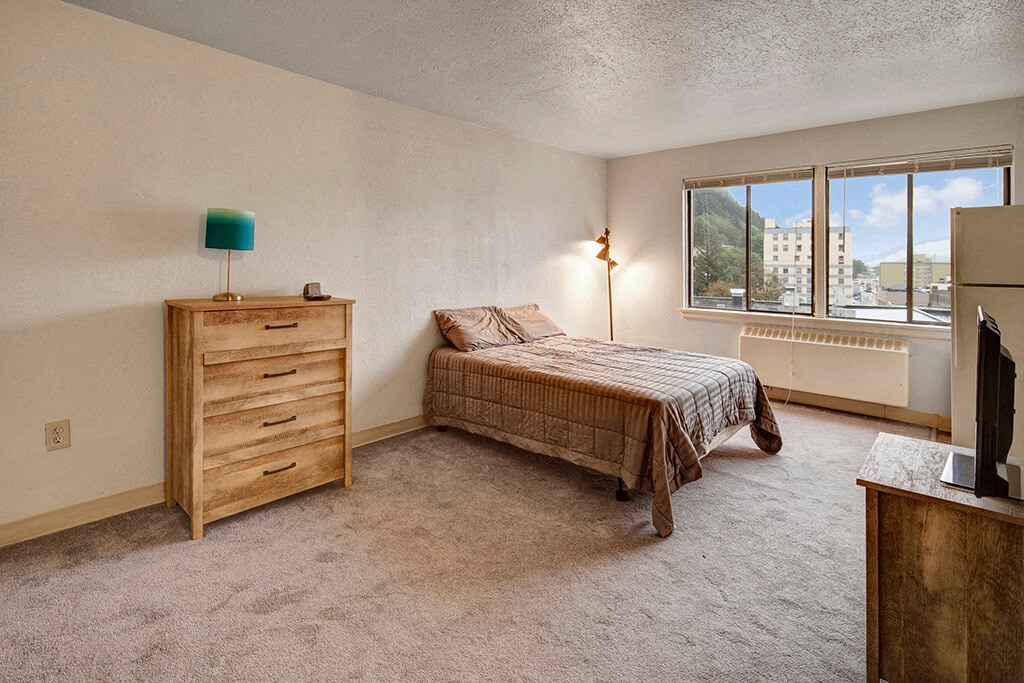 A simple, cozy bedroom here at Mendenhall Tower Apartments with a single bed by the window, draped in a brown plaid blanket. A wooden dresser with a blue lamp sits nearby. Soft lighting creates a calm ambiance.
