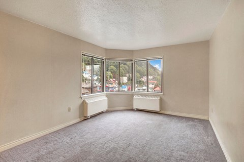 Mendenhall Tower Apartments - Living Room Living room here at Mendenhall Tower Apartments with beige walls and gray carpet, featuring a bay window. The window offers a view of colorful houses and lush green hills under a blue sky.
