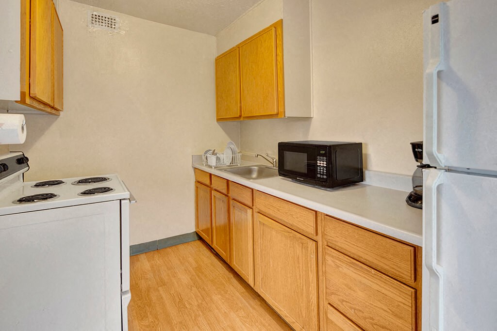 Compact kitchen here at Mendenhall Tower Apartments with light wood cabinets, white appliances, and a microwave. Includes an electric stovetop, sink with dish rack, and a coffee maker.