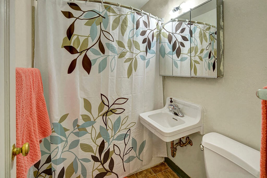 Bathroom here at Mendenhall Tower Apartements with a leafy pattern shower curtain, white sink, and mirror. A coral towel hangs nearby, creating a calm, fresh atmosphere.