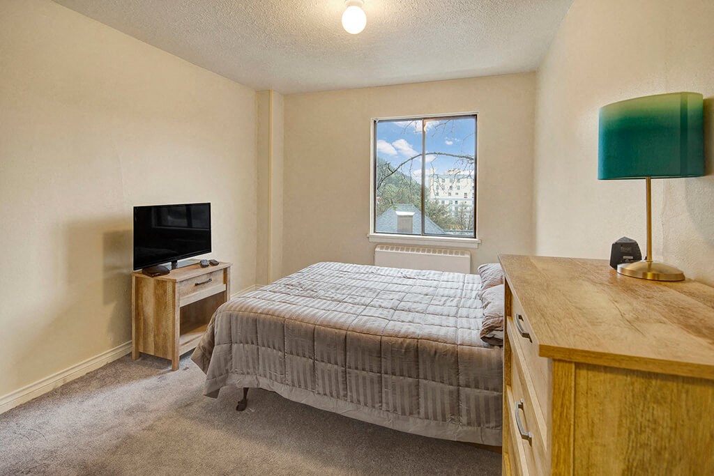 A cozy bedroom here at Mendenhall Tower Apartment featuring a wooden bed with a gray quilt, a small TV on a stand, a dresser with a teal lamp, and a window with a view of trees.