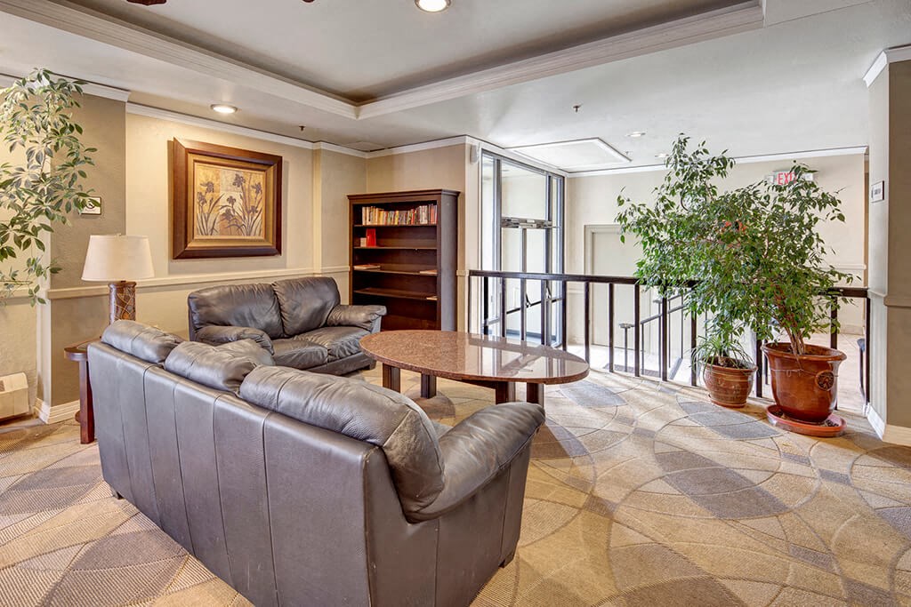 Cozy lounge area here at Mendenhall Tower Apartments with two gray leather sofas facing a round, wooden coffee table. A bookshelf, framed artwork, and large potted plants enhance the warm atmosphere.