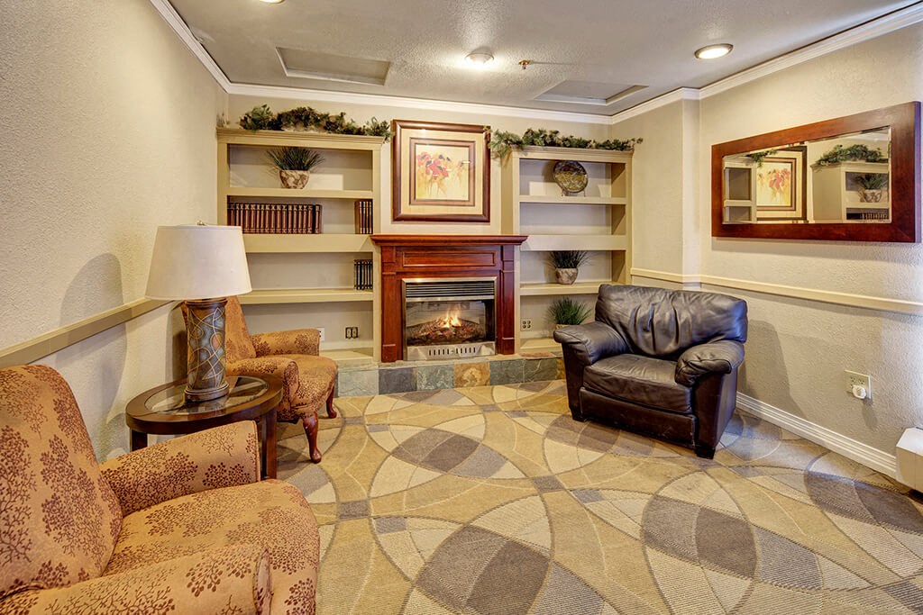 Cozy lounge area here at Mendenhall Tower Apartments with a fireplace, flanked by a brown leather chair and floral-patterned sofas. Bookshelves, a framed painting, and a mirror enhance warmth.
