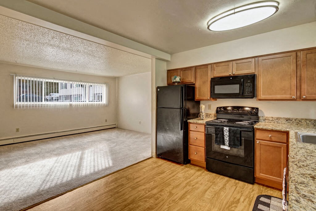 Cozy apartment interior here at Montclair Apartments with an open layout. The kitchen has wooden cabinets, black appliances, and a granite countertop. Large window with blinds. Bright and inviting.