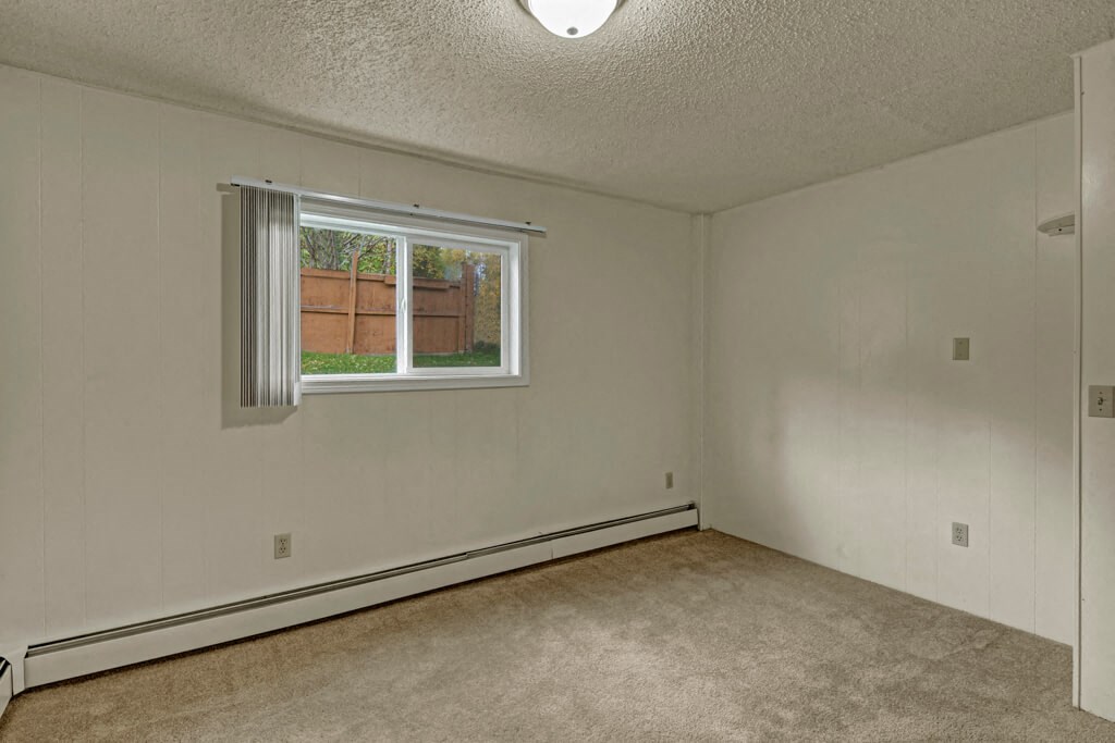 Empty bedroom here at Montclair Apartments with beige carpet and off-white walls, featuring a single window with vertical blinds and a view of a wooden fence and greenery outside. Neutral and calm atmosphere.