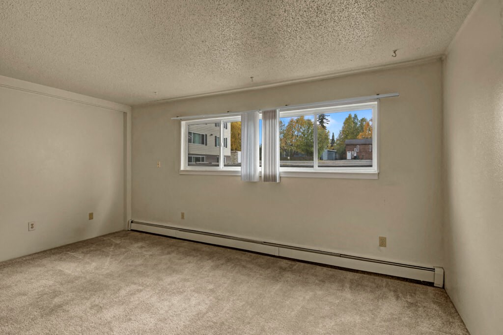 Empty bedroom here at Montclair Apartments with light beige walls and carpet, large window with vertical blinds showing trees and a building outside. Calm, neutral atmosphere.