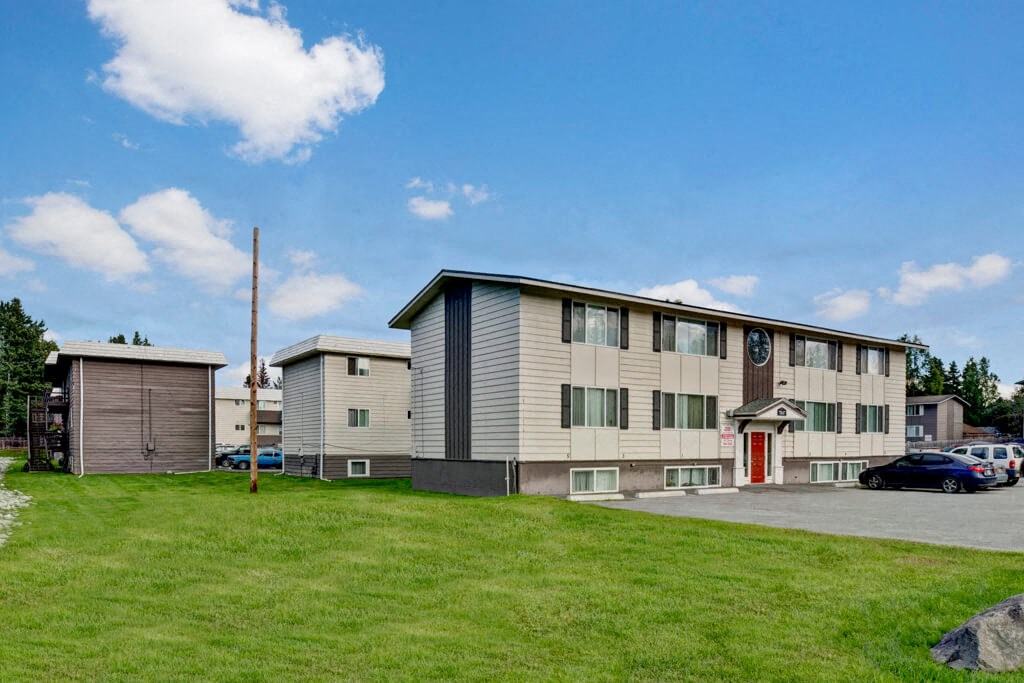 Two-story Montclair apartment building with white siding and red door set against a blue sky with fluffy clouds. Surrounding green lawn adds a serene touch.