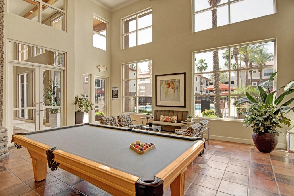 Bright recreation room here at Mozaic at Steele Park Apartments with a pool table, modern seating, large potted plants, and expansive windows showcasing a view of palm trees and a pool.