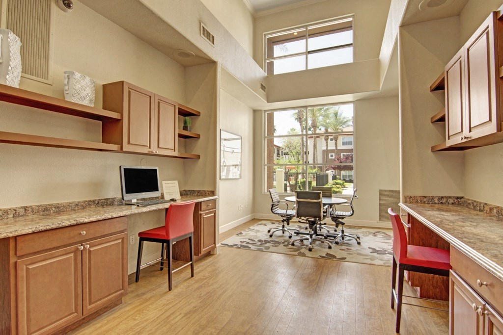 Modern office space here at Mozaic at Steele Park Apartments with wooden floors, large window, two workstations with computers, red chairs, and a round meeting table with rolling chairs. Bright and airy ambiance.