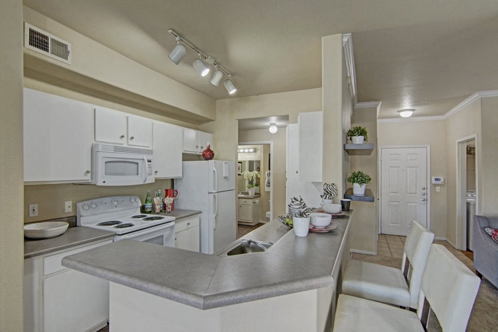 Modern kitchen here at Mozaic at Steele Park Apartments with white cabinets, gray countertops, and bar seating. Bright lighting, decorative plants, and a cozy, welcoming ambiance.