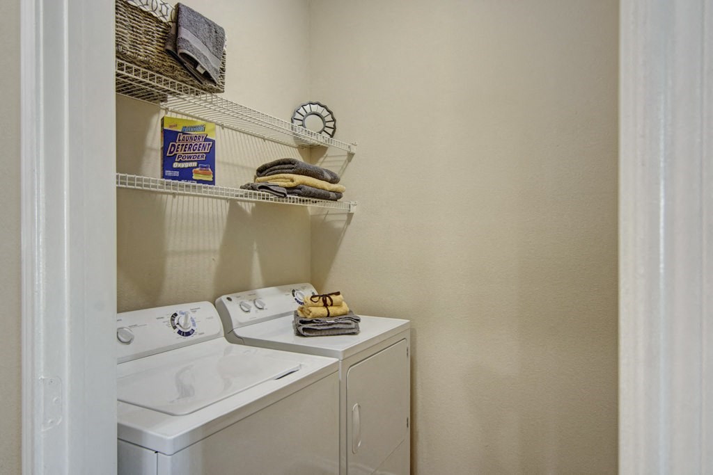 Compact laundry room here at Mozaic at Steele Park Apartments with a washing machine and dryer. Shelves hold detergent, a wicker basket, and neatly folded towels, creating an organized feel.