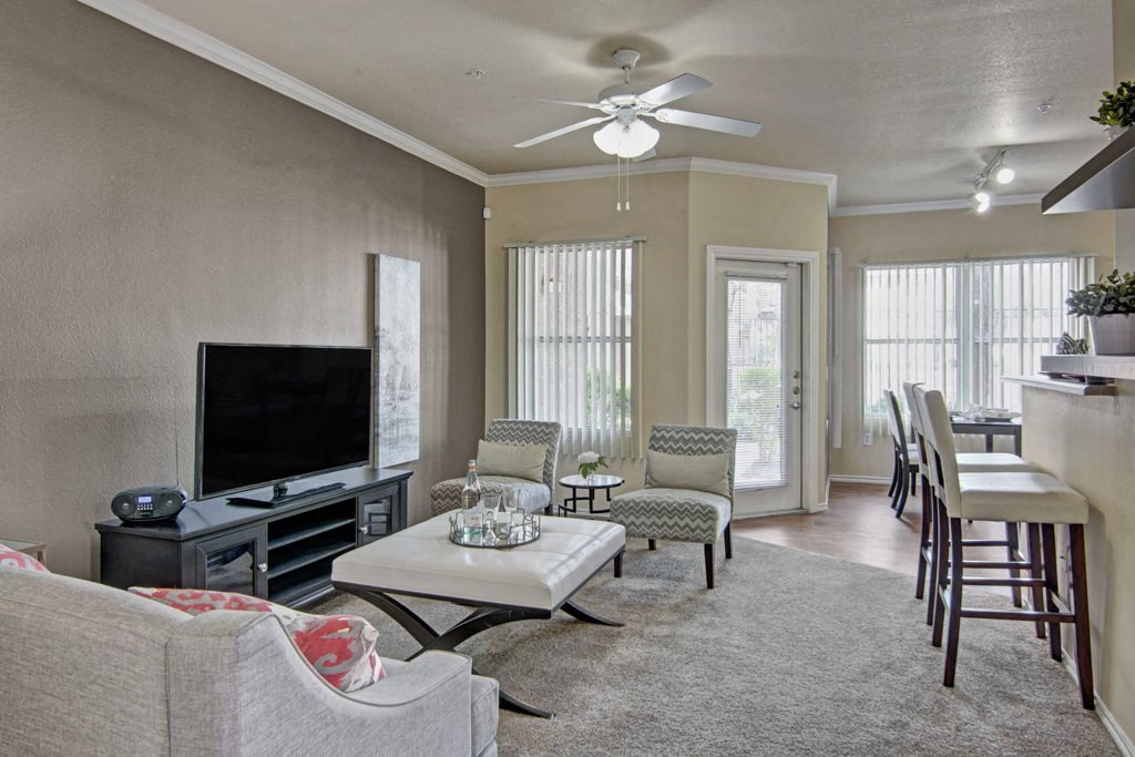 Modern living room here at Mozaic at Steele Park Apartments with neutral tones, featuring a TV on a stand, a gray sofa, patterned chairs, bar stools, and tall windows letting in natural light.