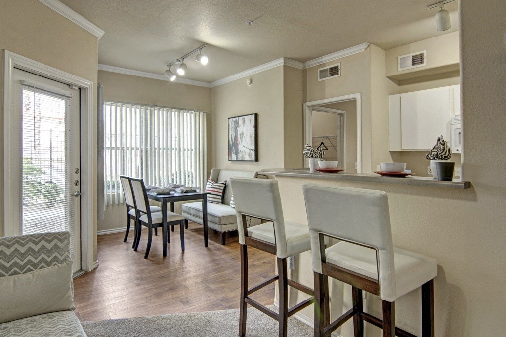 A cozy apartment interior here at Mozaic at Steele Park Apartments with a dining area and kitchen. The dining area has a black table and chairs by large windows. The kitchen features a breakfast bar with high chairs, evoking a warm, inviting atmosphere.