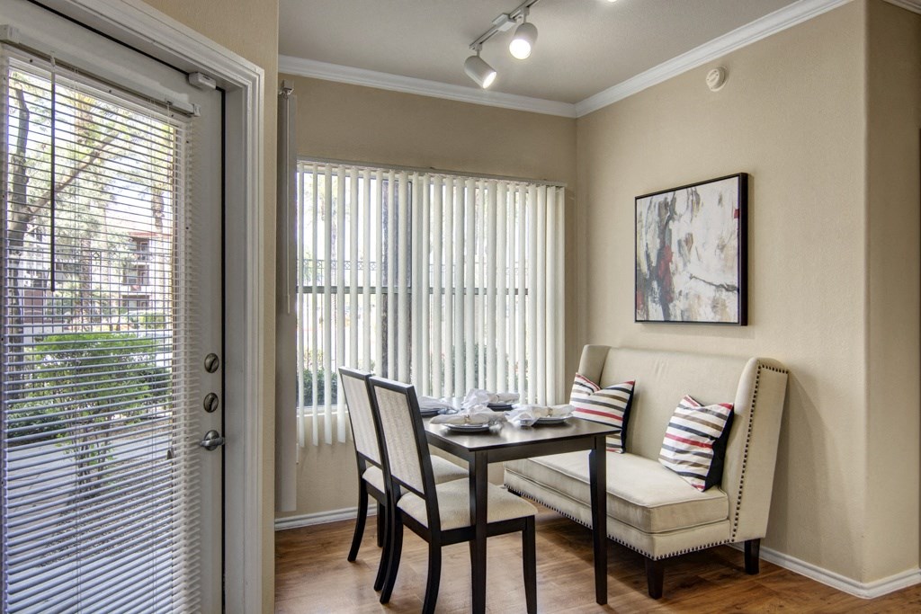 Cozy dining area here at Mozaic at Steele Park Apartments with a beige bench and chairs at a wooden table set for four. Large windows with vertical blinds let in natural light. Abstract art on the wall.