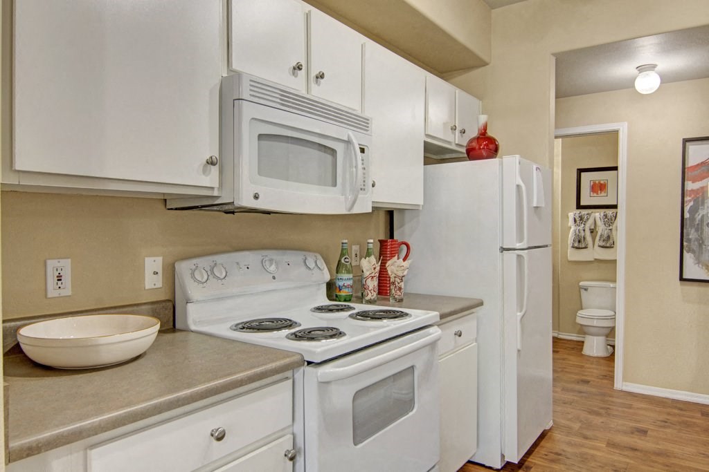 A cozy kitchen here at Mozaic at Steele Park with white cabinets, stove, and microwave. A red vase sits atop the fridge. A tidy bathroom with framed art is visible in the background.