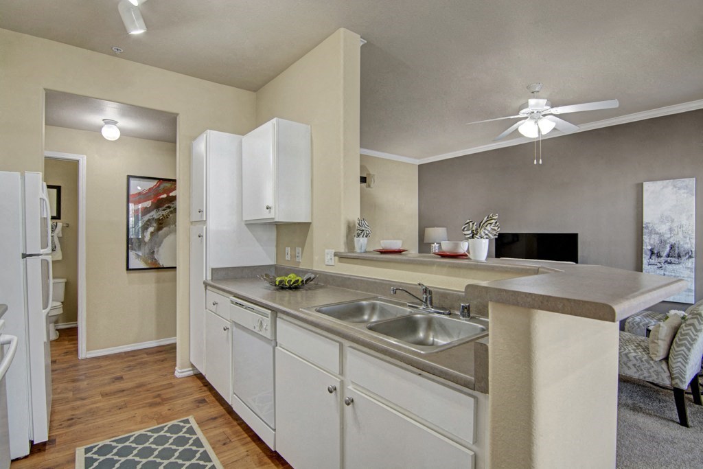 Modern kitchen view here at Mozaic at Steele Park Apartments with beige walls and wood flooring. White cabinets, stainless steel sink, and neutral decor including a fan and art piece. Cozy and inviting.