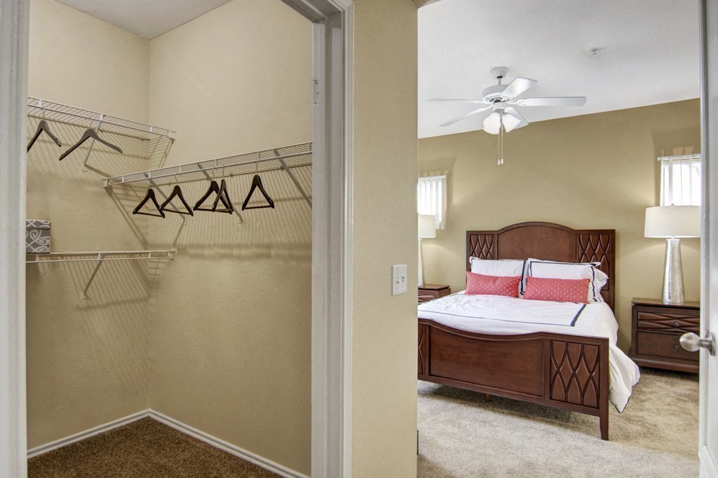 Bedroom view here at Mozaic at Steele Park Apartments from a closet entrance, featuring empty hangers on wire shelves. Warm tones, neatly made bed with red pillows, and serene ambiance.