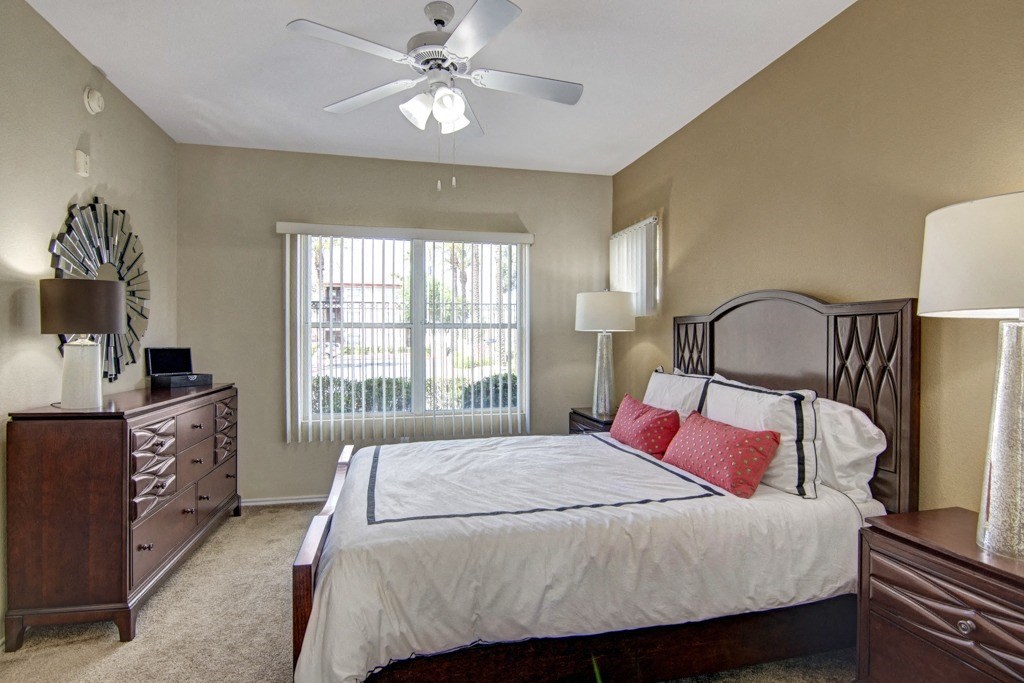 Cozy bedroom here at Mozaic at Steele Park Apartments with a large bed, white linens, and red accent pillows. A ceiling fan hangs above. Wooden furniture and large window create a warm atmosphere.