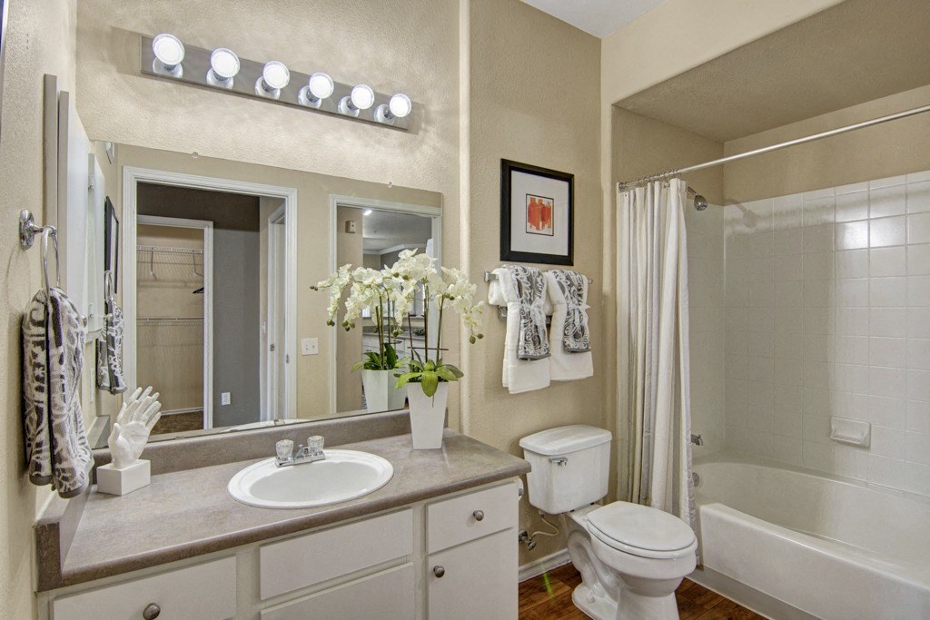 Bright, modern bathroom here at Mozaic at Steele Park Apartments with large mirror, countertop sink, white cabinets, and vase of flowers. Towel racks, framed art, shower, and toilet included.