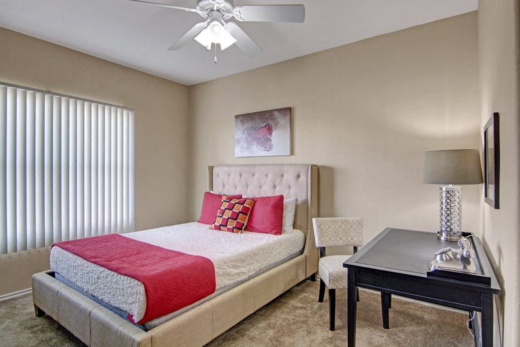A cozy bedroom here at Mozaic at Steele Park Apartments with a beige tufted bed, white and red bedding, and a checkered chair. A black desk with a lamp and modern decor adds elegance.