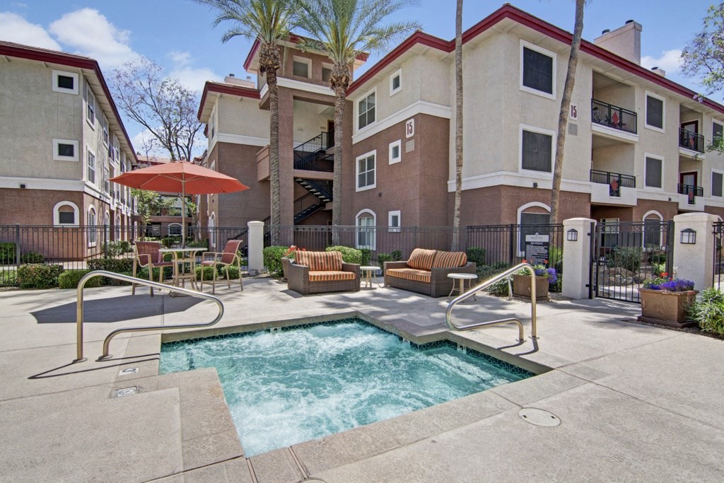 Sunny courtyard with bubbling hot tub, surrounded by metal railing. Two-story beige Mozaic at Steele Park Apartment building, palm trees, and patio chairs create a relaxing vibe.