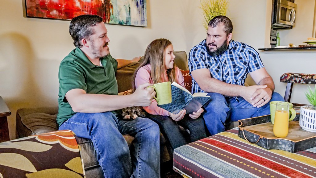Two dads sitting on the couch reading with their daughter in their living room at Bristol Square Apartments