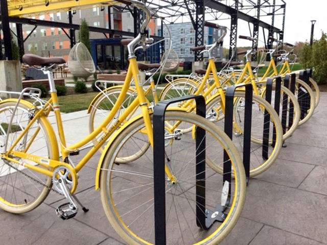 A row of bright yellow bicycles is lined up neatly at a bike rack outdoors, with an industrial building in the background, outside Stitchweld Apartments.