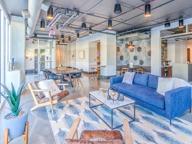 Modern lounge with a stylish blue sofa, geometric rug, and wooden chairs. Exposed ceiling pipes and patterned walls create a chic, inviting atmosphere at Stitchweld Apartments.