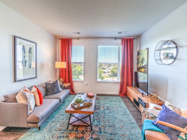A bright, cozy living room with a gray couch, colorful cushions, wooden coffee table, large rug, vibrant red curtains, and a flat-screen TV at Stitchweld Apartments.