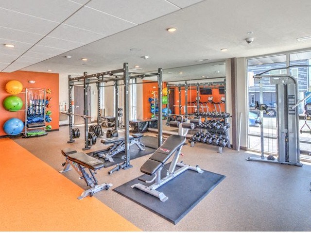 Modern gym with weightlifting equipment, exercise balls, and mirrors. Bright and spacious with warm-toned walls, creating an inviting atmosphere at Stitchweld Apartments.