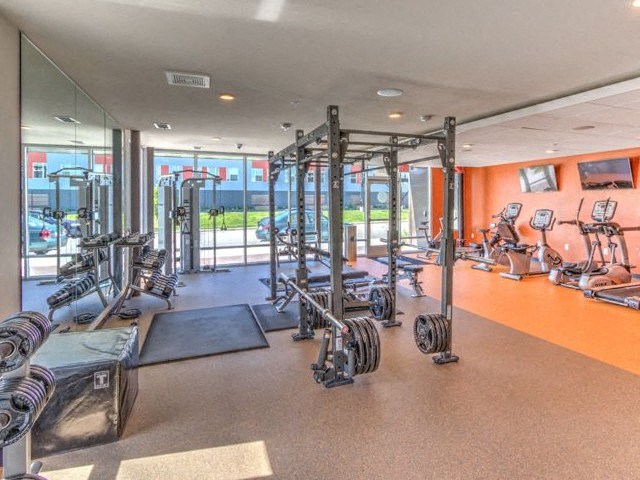 Bright gym with large windows, weightlifting equipment, and exercise machines. Orange accent wall adds warmth at Stitchweld Apartments.