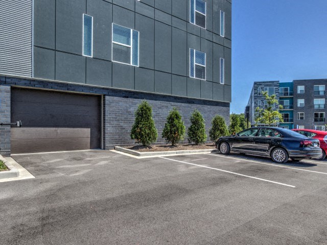 Parking lot with gray building on the left, featuring small trees lined along the side. A parked black car is visible under a clear blue sky at Stitchweld Apartments.